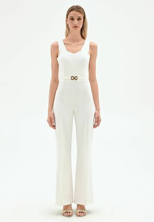 Donna con capelli biondi lisci che indossa una tuta bianca senza maniche con dettaglio cintura e tacchi dorati con cinturini, in piedi su sfondo bianco.