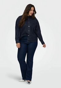 Mørkeblå denim skjorte med knapper og et lyst, spraglete mønster, brukt sammen med mørkeblå vide jeans og sølvfargede sko med spiss tå.