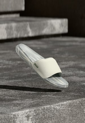 Sandalia tipo chancla White Reef blanca suspendida sobre escalones de hormigón gris texturizado con luz natural suave, mostrando el logo lateral y detalles de la suela.