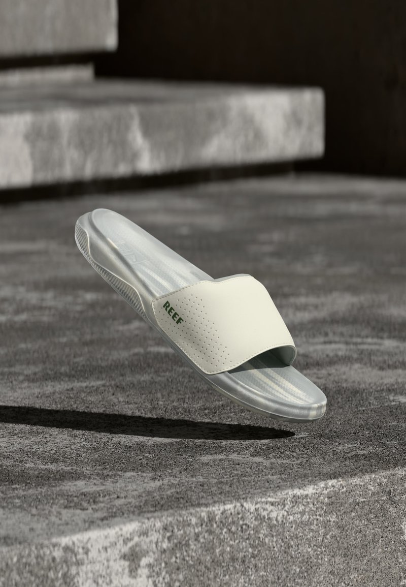 Sandale slide White Reef blanche suspendue au-dessus de marches en béton gris texturé sous une lumière naturelle douce, montrant le logo latéral et les détails de la semelle.