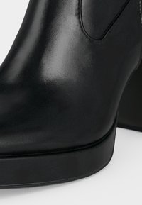 Gros plan d’une botte cheville en cuir noir avec un bout arrondi et un petit talon bloc sur fond blanc.