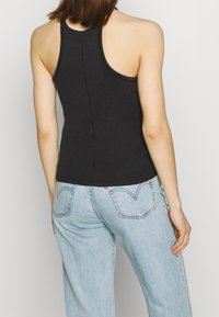 Person, der ein schwarzes ärmelloses Racerback-Top und eine hellblaue High-Waist-Jeans trägt, von hinten gegen einen schlichten Hintergrund gezeigt.