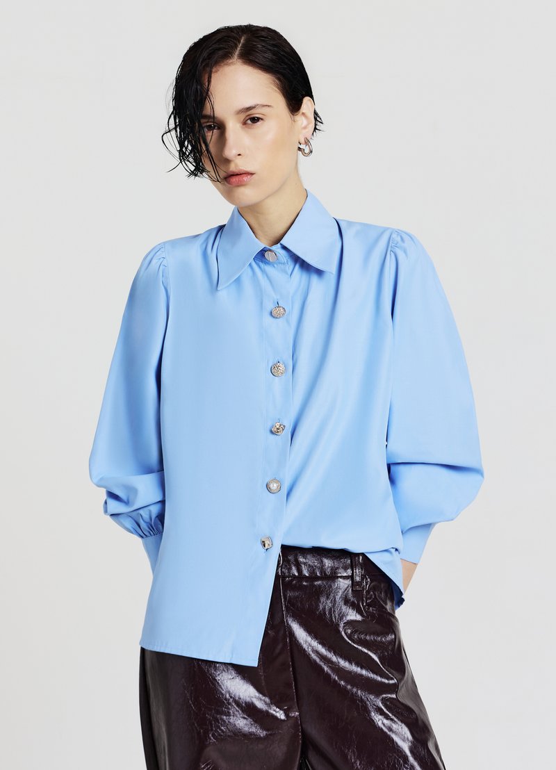 Light blue blouse with a large collar and puffed sleeves, featuring silver buttons down the front. Paired with shiny dark brown pants.