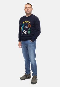 Granatowy sweter z kolorowym haftem "Après Ski", niebieskie dżinsy i szare buty sportowe z czarną podeszwą. Mężczyzna stoi w neutralnej pozycji.