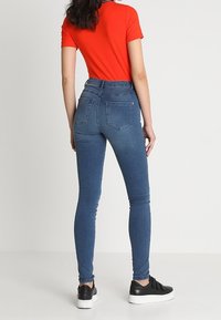 Woman wearing fitted blue jeans, red short-sleeve top, and black sneakers, standing with back facing the camera against a plain background.