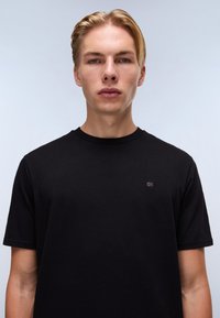 Fekete, rövid ujjú póló, pamutból készült, kerek nyakkivágással és kis logóval az elején, sima textúrával.