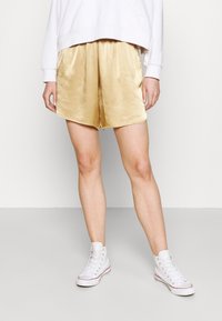 Satinhose in Gold mit elastischem Bund, glatt in der Textur und locker geschnitten, kombiniert mit weißen High-Top-Sneakern.