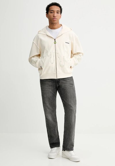 Felpa crema con zip e maniche oversize, indossata sopra una camicia bianca, abbinata a jeans grigi e sneakers bianche.