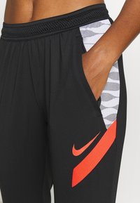 Zwarte sportbroeken met een mesh tailleband, zijzakken met een grijs en wit patroon, en een felle rode Nike-logo accent.