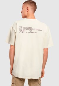 Overdimensioneret creme t-shirt med et trykt grafisk motiv på ryggen, der inkluderer tekst og et blomsterikon, korte ærmer og en afslappet pasform.