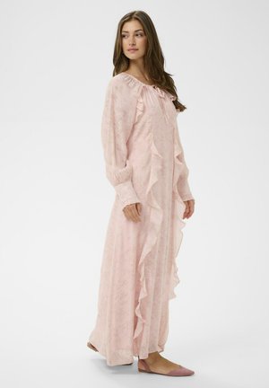 Nő áll, hosszú rózsaszín virágos ruhát visel fodros részletekkel és hosszú ujjakkal, rózsaszín lapos cipővel párosítva, oldalra néz.