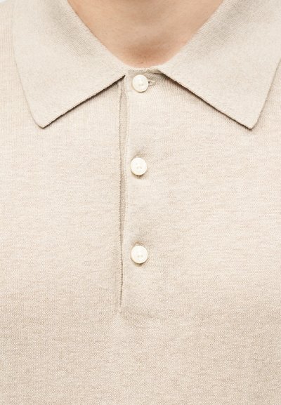 Μπεζ πλεκτό πόλο πουκάμισο με μικρό γιακά και τρία λευκά κουμπιά, φορεμένο από άτομο που φαίνεται από το λαιμό μέχρι το πάνω μέρος του στήθους.