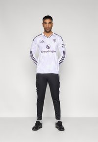 Camisa de futebol branca de manga comprida com detalhes roxos e padrões geométricos, combinada com calças pretas com riscas brancas nas laterais.