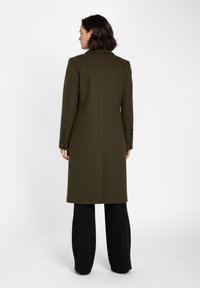 Olivgrön skräddarsydd kappa med en strukturerad design, axelkuddar och fem svarta knappar på ärmarna. Bärs över svarta byxor.