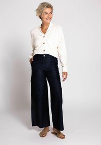 Cremefarbener, verkürzter Cardigan mit Knöpfen, kombiniert mit weiten Cargo-Hosen aus dunklem Denim mit Seitentaschen. Einfache Sandalen runden das Outfit ab.