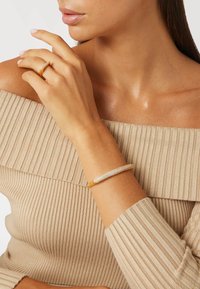 Goldfarbener Armreifen mit kunstvollen Kristallverzierungen, glatter Oberfläche und länglich-ovalem Design, getragen auf einem strukturierten beigen Oberteil.