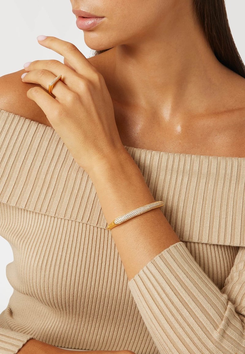 Goldfarbener Armreifen mit kunstvollen Kristallverzierungen, glatter Oberfläche und länglich-ovalem Design, getragen auf einem strukturierten beigen Oberteil.