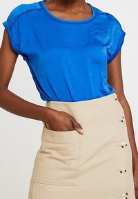 Blusa azul de manga corta con una textura suave y un escote redondo, combinada con una falda beige que presenta un bolsillo lateral y detalles de botones.
