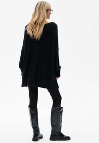 Zwarte oversized gebreide trui met een losse fit, brede mouwen en een geribbelde zoom, gecombineerd met zwarte skinny broeken en hoge zwarte leren laarzen.
