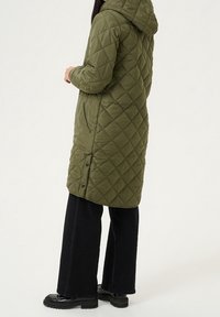 Long, olive green quilted coat with hood, featuring side pockets and snap closures. Worn over black wide-leg pants and chunky black boots.