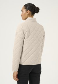 Veste matelassée beige clair avec un col montant, des manches longues et un motif en losanges texturé. Sans fermetures visibles. Porté avec un pantalon noir.