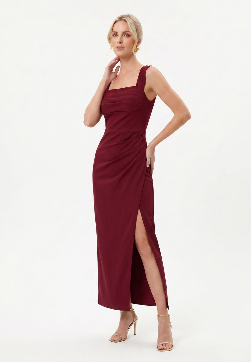 Femme aux cheveux blonds portant une robe bordeaux sans manches avec une fente latérale et des sandales à talons hauts beige, debout sur fond blanc.