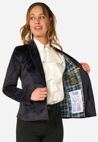 Mörk sammetblazer med figursydd passform, spetsiga slag och satinliknande textur. Insidan har ett rutigt mönster med tryckta etiketter. Vit skjorta med volanger under.