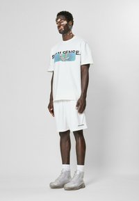 T-shirt blanche avec impression graphique et texte sur le devant, assortie à un short en maille blanche. De grosses baskets grises complètent la tenue.