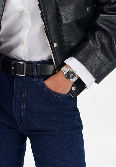 Czarna skórzana kurtka, biała koszula i ciemnoniebieskie dżinsy. Srebrny zegarek z czarną tarczą, teksturowanym paskiem i metalową klamrą.