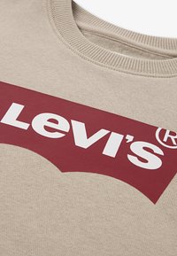 Beige sweatshirt med en framträdande röd Levi's-logotyp i vitt, har rund halsringning och texturerade tygdetaljer.