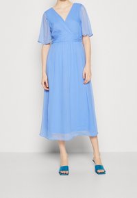 Robe bleu clair mi-longue avec un design cache-cœur, manches transparentes et taille cintrée. Associée à des mules bleu ouvertes à l'avant. Texture lisse.