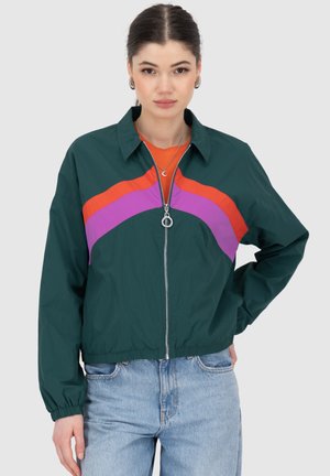 Veste coupe-vent vert foncé avec des rayures horizontales orange et violettes, dotée d'un col, d'une fermeture éclair frontale et de poignets élastiques. Associée à un jean clair.