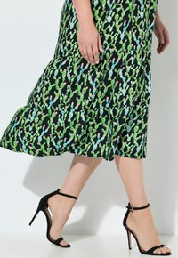 Schwarzes Kleid mit grünem und pinkem Kaktusdrucken; gestufter Schnitt. Das Model trägt schwarze High Heels mit Knöchelriemen.