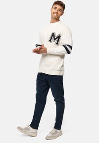 Hvid strikket cardigan med et stort marineblåt "M" på venstre bryst, med marineblå stribede detaljer på ærmerne, parret med mørke jeans og beige sneakers.
