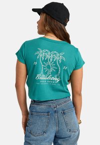 Teal cropped t-shirt met ribbeltextuur; witte bedrukte palmbomen en tekst op de achterkant. We ingevuld met hooggeprijsde blauwe spijkerbroeken. Zwarte pet op.