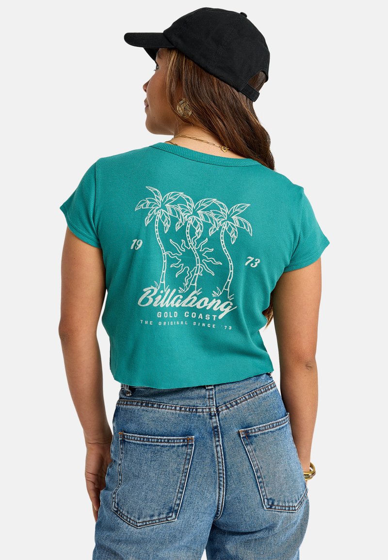 Teal cropped t-shirt met ribbeltextuur; witte bedrukte palmbomen en tekst op de achterkant. We ingevuld met hooggeprijsde blauwe spijkerbroeken. Zwarte pet op.