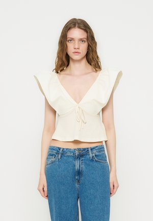 Jeune femme portant un crop top crème à volants avec un décolleté plongeant en V et un jean en denim bleu taille haute, debout devant un fond uni.