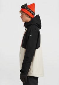 Personne jeune portant une veste à capuche noire et beige, un bonnet orange avec des lunettes de ski, et des gants noirs, debout de côté contre un fond uni.