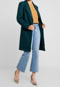 Kvinna i mörkgrön kappa över en orange tröja, ljusblå korta utsvängda jeans och högklackade skor med ormskinnsprint som står på vit golv.