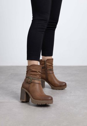 Botas de cuero marrón hasta el tobillo con un diseño arrugado, acentos metálicos, correa ajustable y un tacón grueso. Suela texturizada para mayor agarre.