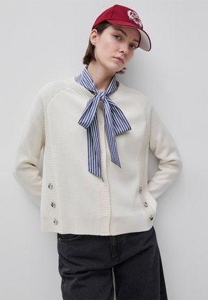 Junge Person trägt eine rote Kappe, einen cremefarbenen Knopfcardigan, einen blau-weiß gestreiften Halsschal und schwarze Hosen, steht vor einem hellen Hintergrund.