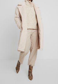 Person som bär en beige konstpälsjacka, en kraftig stickad tröja, ljusbeiga byxor och spetsiga ankelstövlar i ormskinnsmönster, st år mot en enfärgad bakgrund.