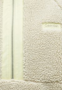 Μπεζ φλις μπουφάν με υφή, με κεντρικό φερμουάρ σε ασημί μεταλλικό υλικό και τμήμα με πάνελ σε ανοιχτό κίτρινο χρώμα. Παρουσιάζεται το λογότυπο της Calvin Klein.