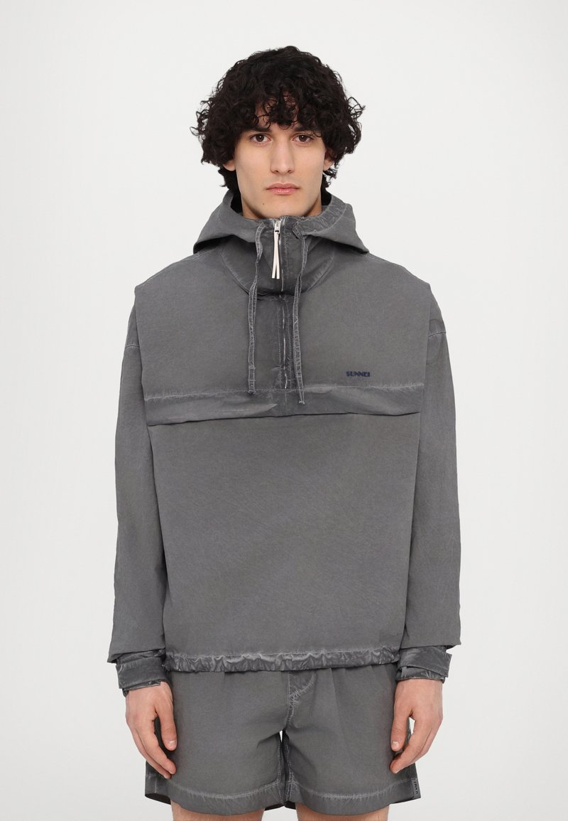 Pullover grigio con colletto alto e tasca frontale, caratterizzato da una finitura testurizzata e cuciture a vista. Abbinato a pantaloni corti coordinati.