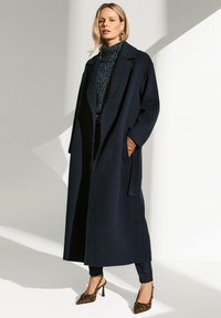 Langer Marineblauer Wollmantel mit übergroßen Revers und seitlichen Taschen, kombiniert mit einem strukturierten blauen Rollkragenpullover und dunklen Hosen.