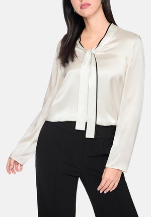Vrouw draagt een crèmekleurige satijnen blouse met lange mouwen en een gestrikte halslijn, gecombineerd met hoog getailleerde zwarte broek tegen een effen achtergrond.