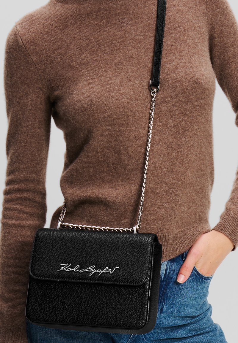 Borsa a tracolla in pelle nera con finitura testurizzata, tracolla in catena argentata e logo ricamato. Indossata sopra un maglione marrone e jeans blu.
