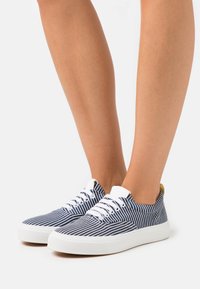 Modré a bílé pruhované plátěné tenisky s bílými gumovými podrážkami, plochými tkaničkami a zlatými detaily na patě. Klasický nízký design.