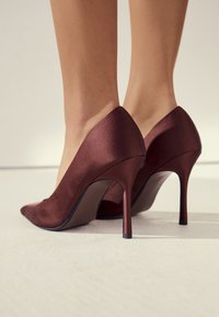 Bordeauxfarvede satin høje hæle med en spids tå og en slank stiletthæl, som har en glat og jævn tekstur.