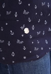 Granatowa koszula z białymi wzorami kotwic i serc, z detalem w postaci srebrnego guzika. Miękki, bawełniany materiał z luźnym krojem.